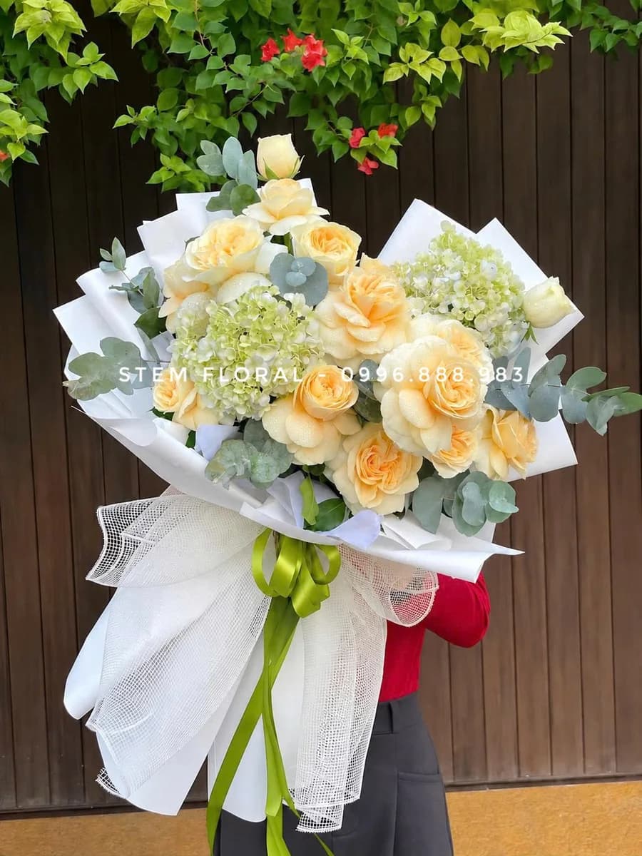 Kem vàng thanh thoát — Hoa hồng vàng kem phối các cụm hoa xanh nhạt và lá xanh, được bó trong giấy trắng mở rộng